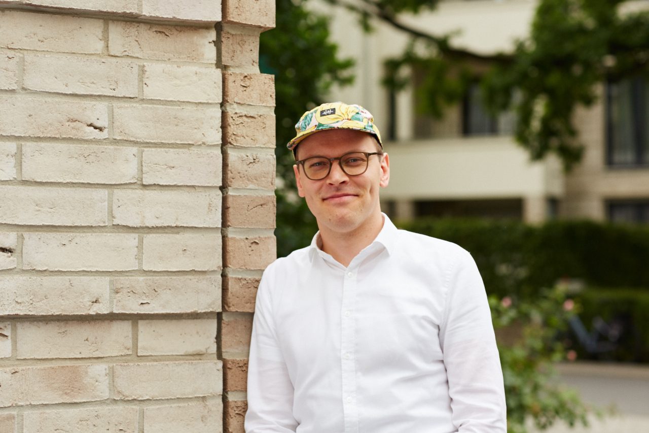 Mann in weißem Hemd und Bananencappy steht an einer Mauer in der Stadt. Im Hintergrund ein Baum verschwommen in der Unschärfentiefe