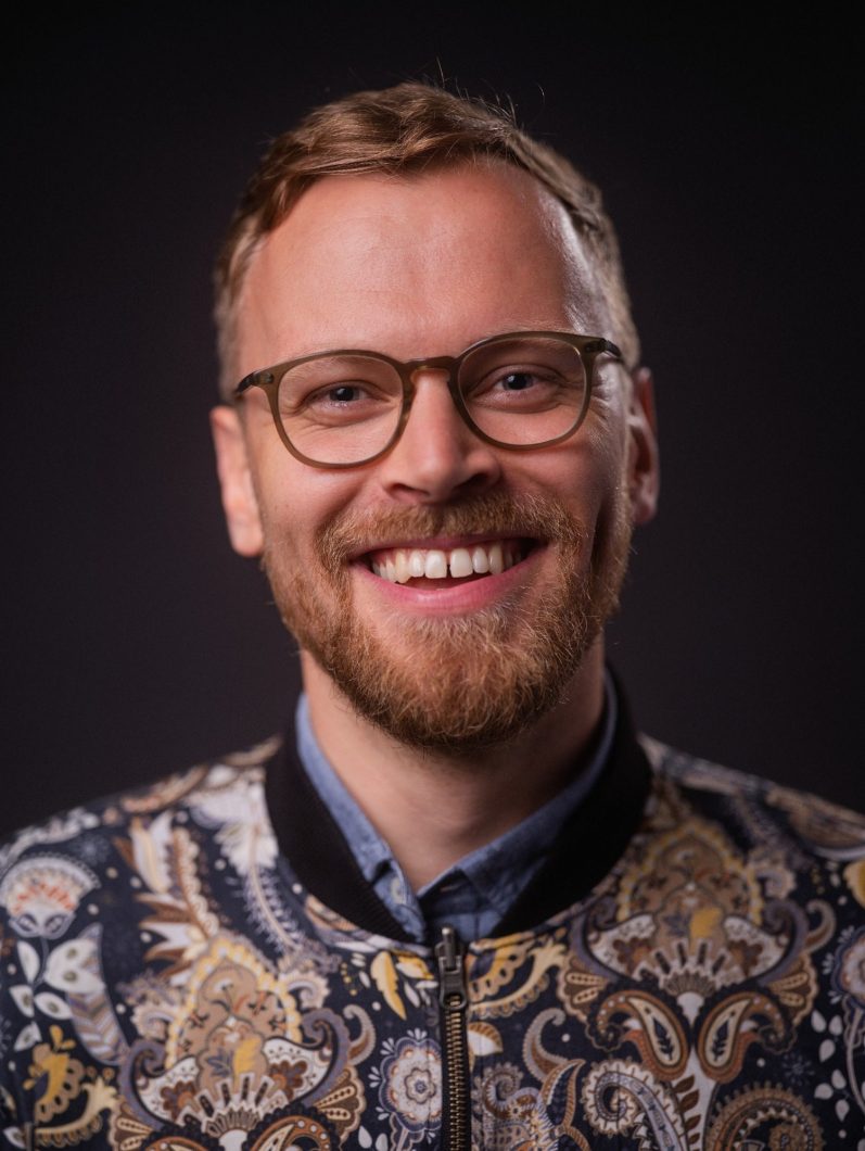 Portrait Robert Marx Mann in weißem Hemd und Bananencappy steht an einer Mauer in der Stadt. Im Hintergrund ein Baum verschwommen in der Unschärfentiefe